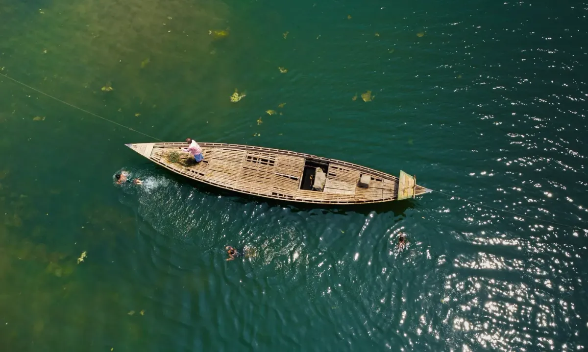 A wooden boat on clear green water at Dawki seen from above, with swimmers beside it