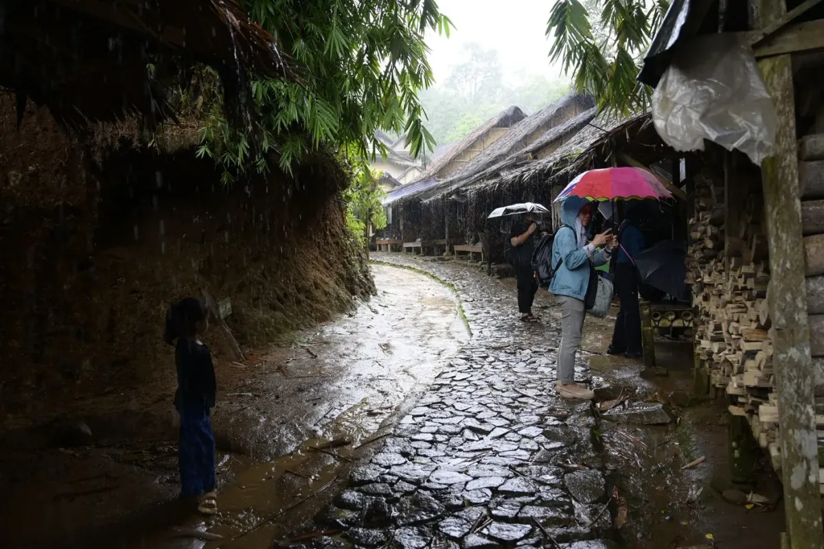 A wet stone lane between wooden houses in a Khasi village, with people under umbrellas